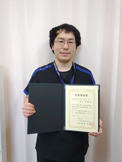 受賞証書を持った男性が微笑んでいる写真。背景は淡い色のカーテン。