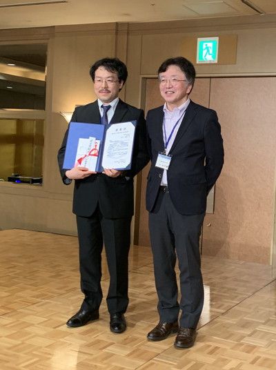 菅谷竜朗先生が第60回日本補体学会学術集会で若手奨励賞を受賞し、賞状を持って笑顔でポーズをとる様子。