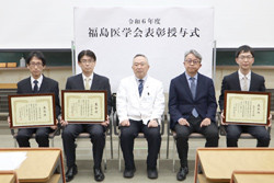 福島県立医科大学の受賞式で、賞状を持つ4人の学生と中央に座る教授の写真。