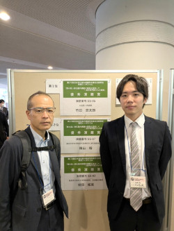 福島県立医科大学の受賞を祝う場面で、二人の男性が掲示板の前に立っている。