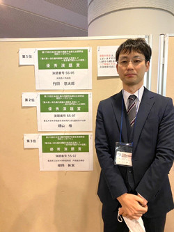 受賞歴を示す掲示板の前に立つ男性。彼はスーツを着ており、表彰状が掲示されています。
