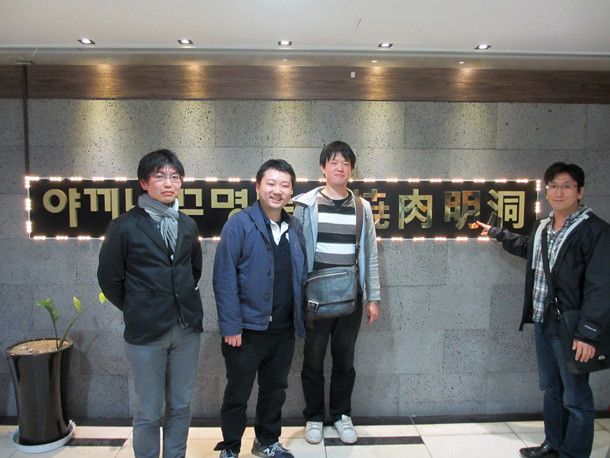 学会参加のために集まった4人の男性が、看板の前でポーズをとっている写真。