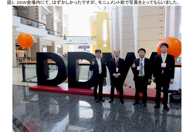 DDW2014の会場で、参加者4名が「DDW」の大きな看板の前で記念撮影をしている様子。