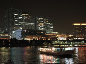 夜の都市景観を背景に、川を行く観光船がライトアップされたビル群の前を進んでいる様子。