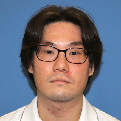 病院助手の関口雅也さんの顔写真。眼鏡をかけた男性で、白いシャツを着用し、青い背景の前に立っています。