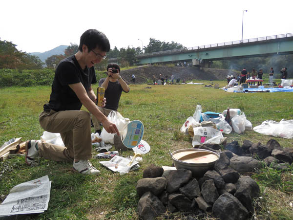 屋外で石のかまどを囲み、男性がうちわで火をあおる様子。