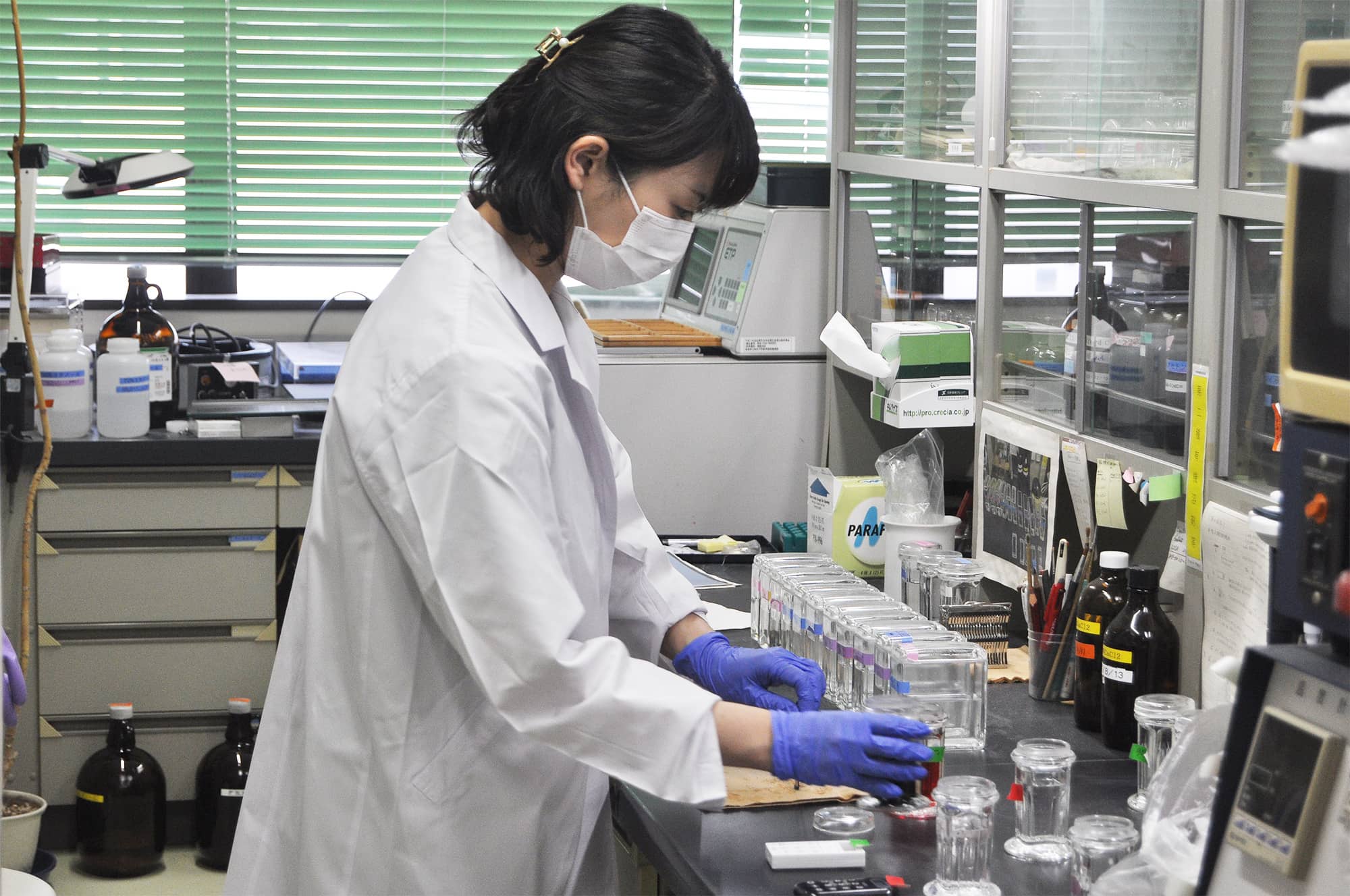 研究室で実験を行う白衣を着た女性が、試験管や容器を扱っている様子。