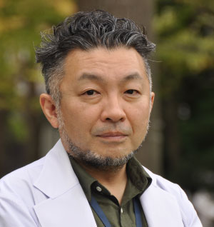 医師の松田希が白衣を着て、自然の中で真剣な表情を浮かべている写真。