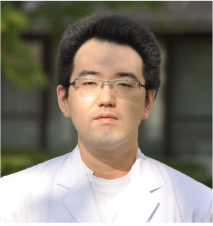 医療白衣を着た阿部暖助手が屋外で真剣な表情で立っている写真。背景には緑の木々が見える。