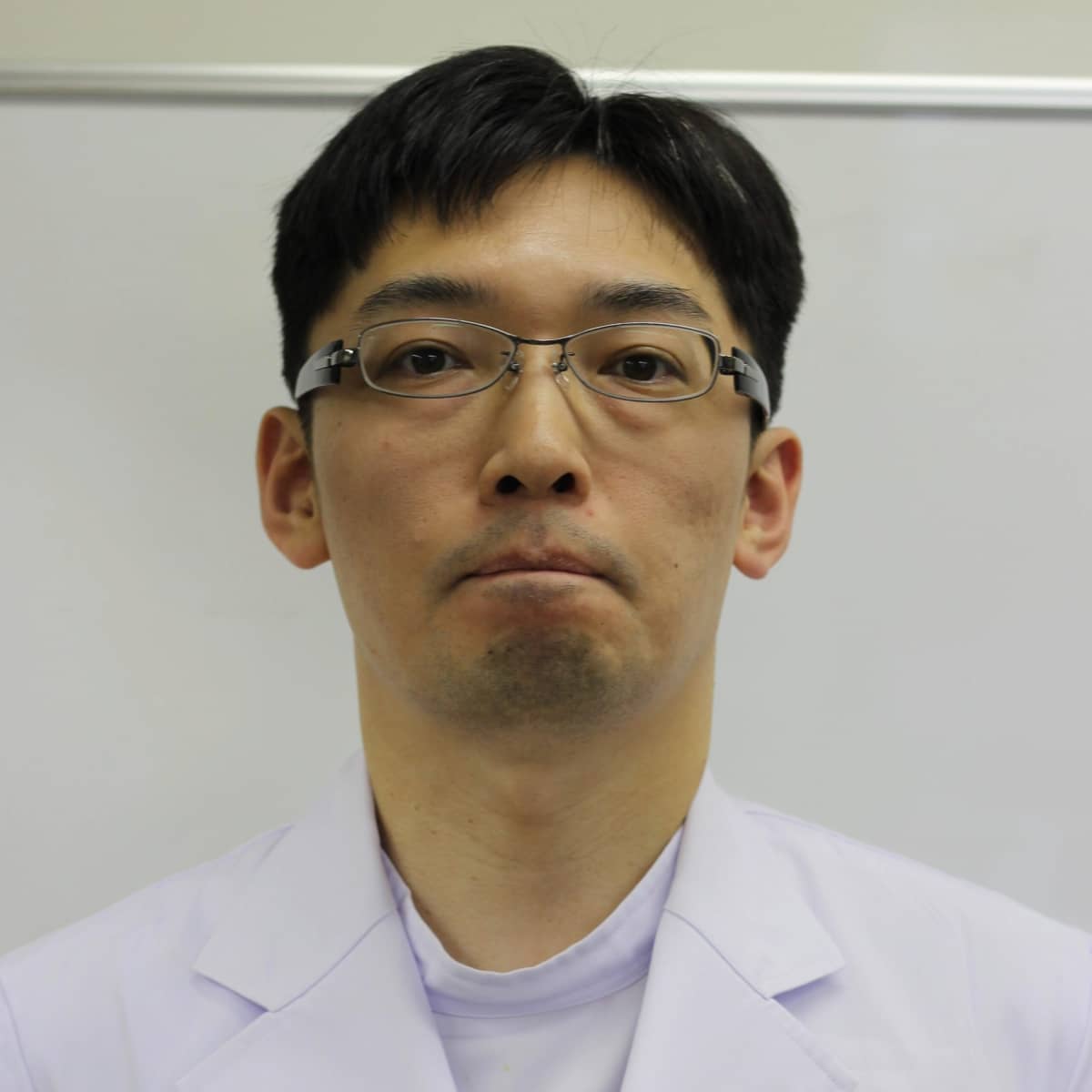 准教授の板垣俊太郎氏が白衣を着て、真剣な表情でカメラを見つめているポートレート。