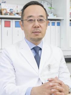 主任教授の佐治重衡が白衣を着て座っている姿。背景には医療関連の書類や道具が見える。