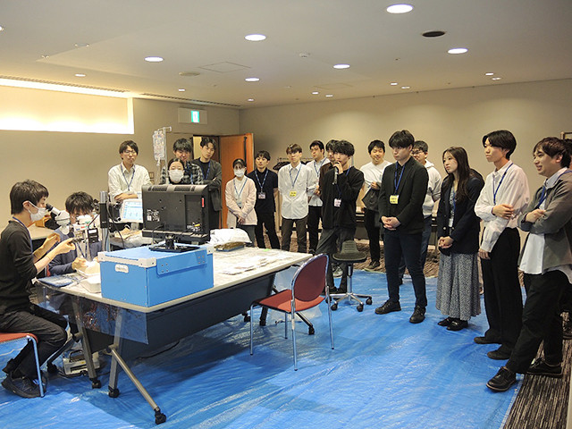 大勢の学生が部屋で医療機器のデモを真剣に見学している場面。医療トレーニングの一環と考えられる。