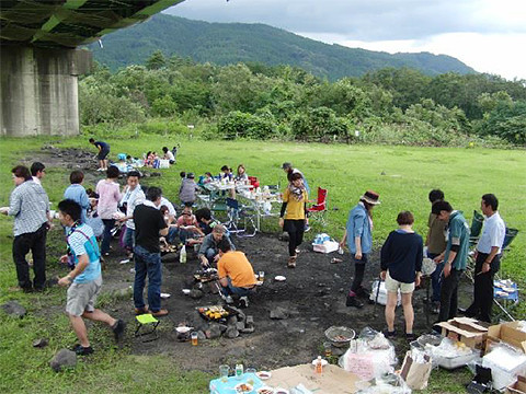 納涼会で楽しむ人々が集まる緑豊かな野外の風景。バーベキューや食事を囲んでいる様子。