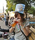 ビールを飲みながら笑顔を見せる女性が、カラフルな帽子をかぶって公園のベンチに座っている様子。周囲には他の人々も見える。
