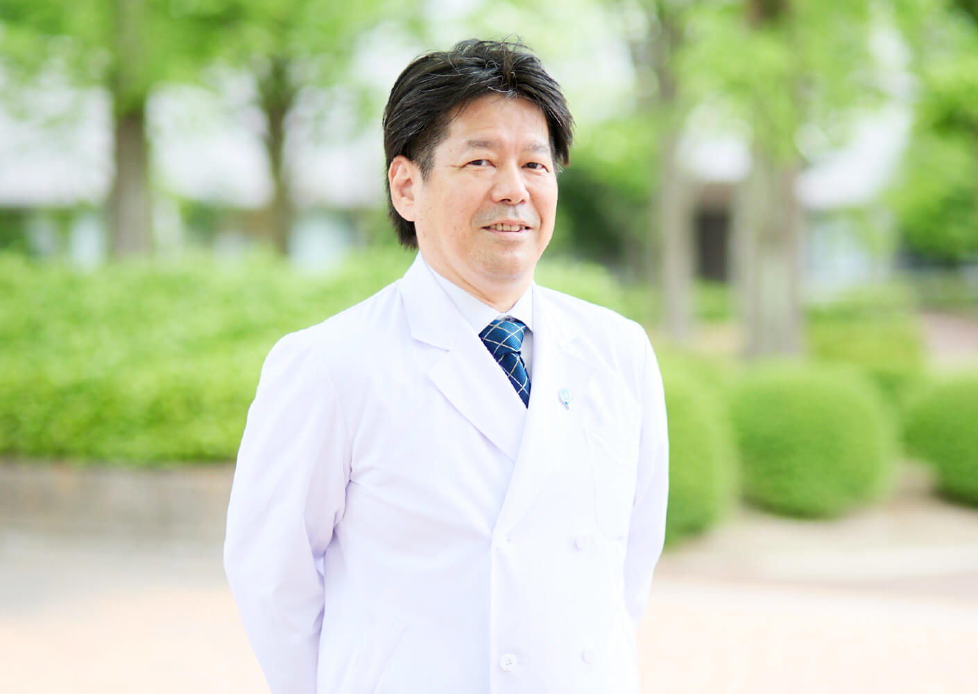 医療白衣を着た男性が緑豊かな公園で微笑んでいる様子。福島県立医科大学の教授と思われる。