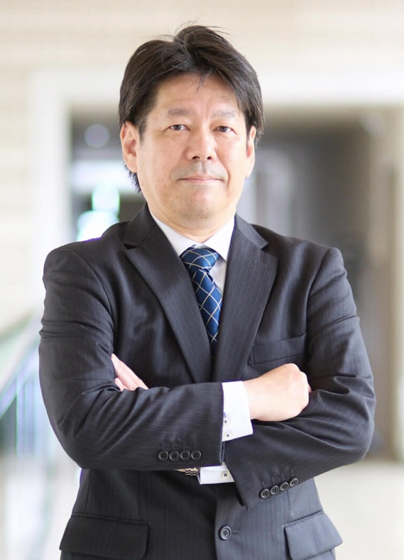 主任教授の鈴木義行がスーツ姿で腕を組み、福島県立医科大学の教室内でポーズを決めている。
