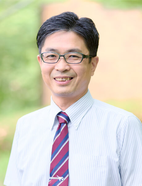 准教授の佐藤久志氏が微笑みながらポーズをとっている写真。背景には自然の緑が広がっている。