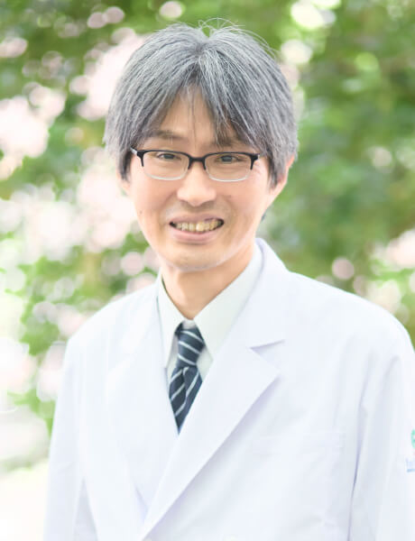 福島県立医科大学の講師、竹本靖氏が白衣を着て微笑んでいる写真。背景には緑の木々が見える。