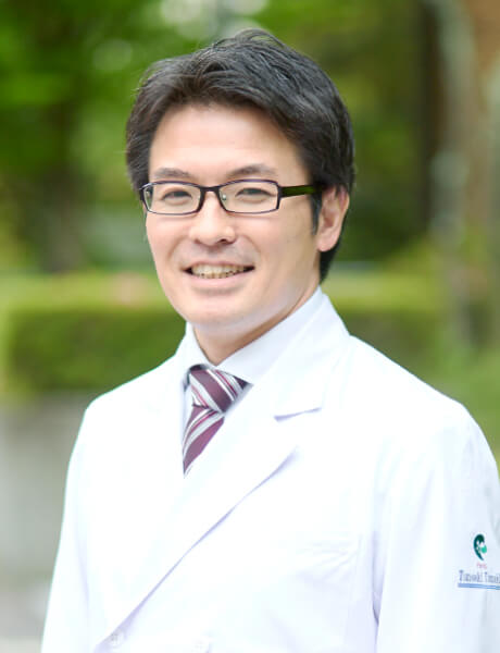 福島県立医科大学の田巻倫明教授が白衣を着て微笑んでいる写真。背景には緑の植物が見える。
