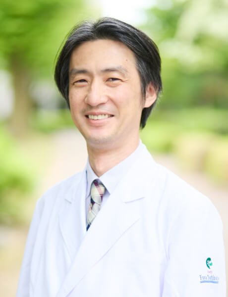 准教授の吉本由哉氏が白衣を着て微笑んでいる写真。背景には緑豊かな公園の風景が広がる。