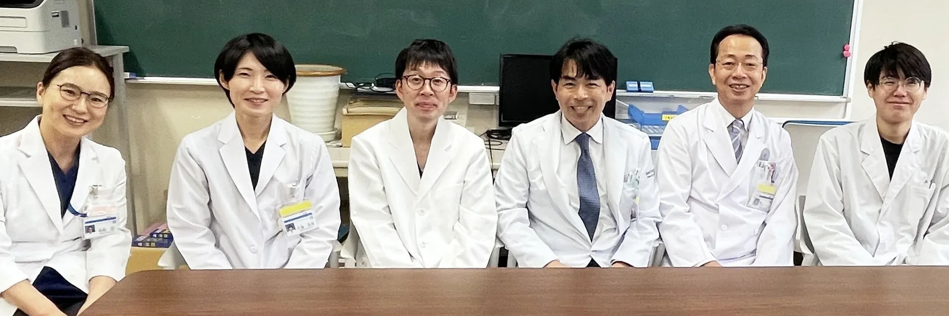 6人の医療関係者が白衣を着て、大学の教室と思われる場所で机越しに笑顔で並んで座っている。バックに黒板がある。