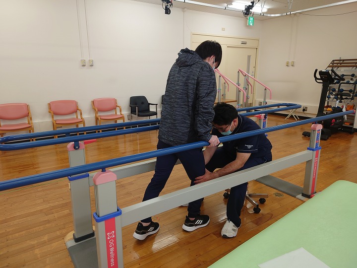 人物が歩行補助バーを使ってリハビリを受けており、トレーナーがその膝を支えてサポートしています。室内は明るく、後方に椅子が並んでいます。