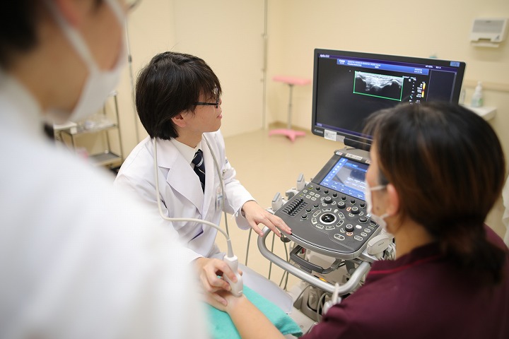 医療用超音波機器を操作する白衣の男性が患者の腕を観察し、他の医療スタッフが見守る様子。