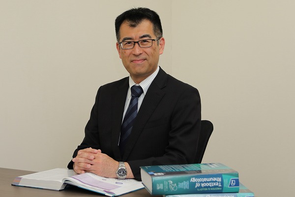 男性がスーツを着て机に座り、開いた本と青い医学書に囲まれています。オフィスのシンプルな内装です。