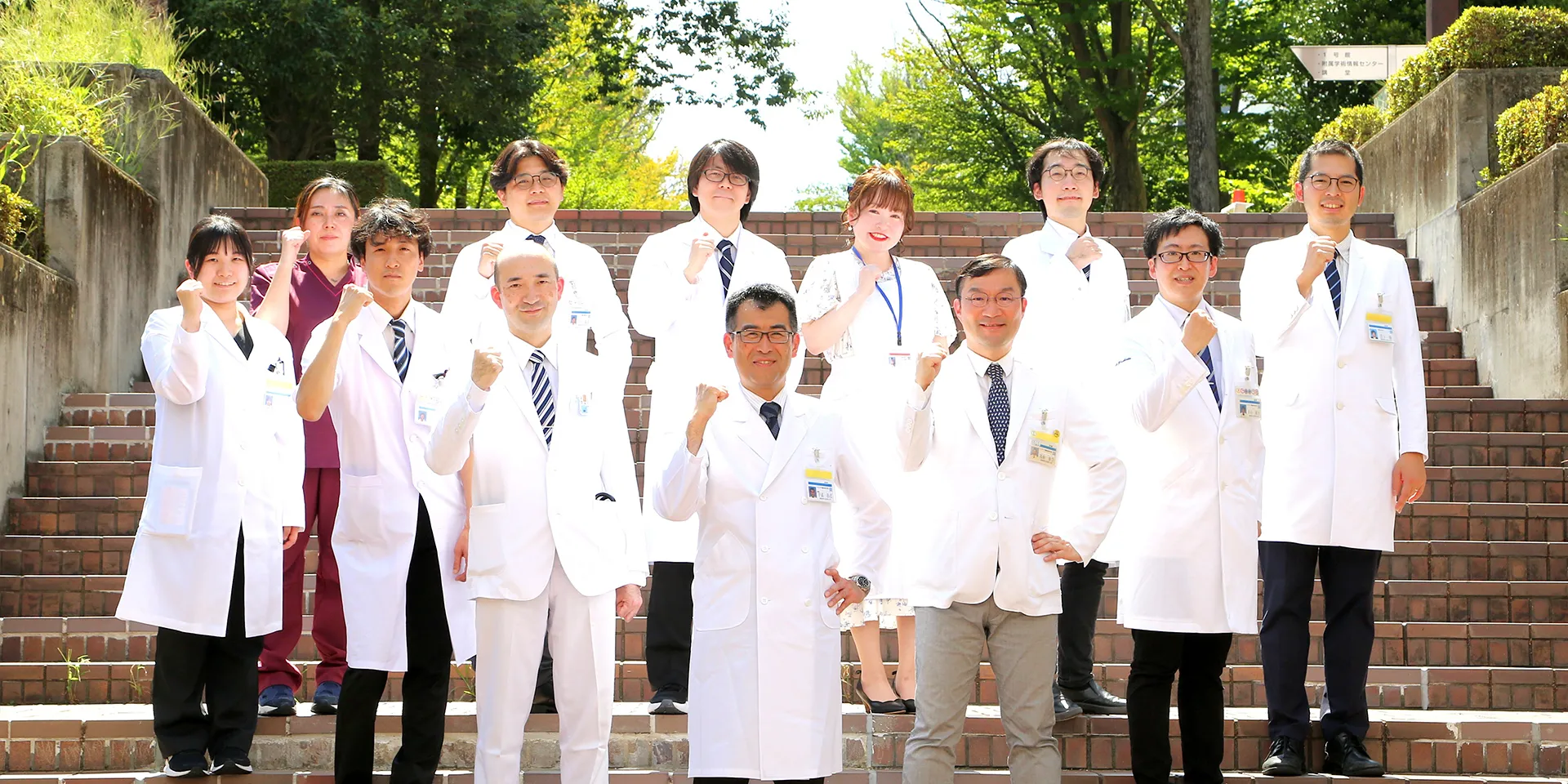 福島県立医科大学の医師たちが白衣を着て集合し、笑顔でカメラに向かっている写真。背景には観葉植物が配置されている。