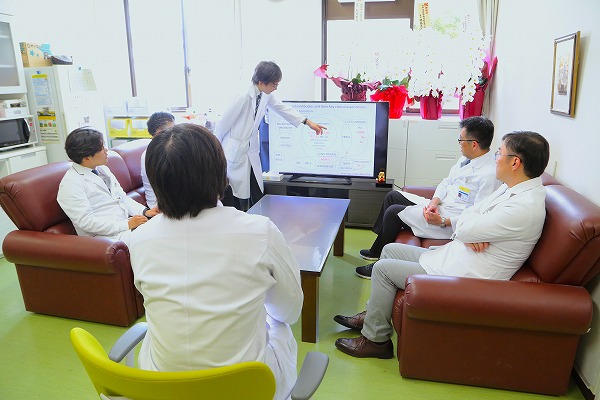 医師たちが会議室でスクリーンを見ながら議論している様子。白衣を着た一人が図を指し説明中。