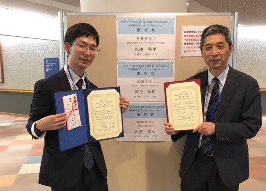 初期臨床研修医の松本聖生先生が、受賞証書を持って笑顔でポーズをとる様子。背景には受賞に関する掲示が見える。