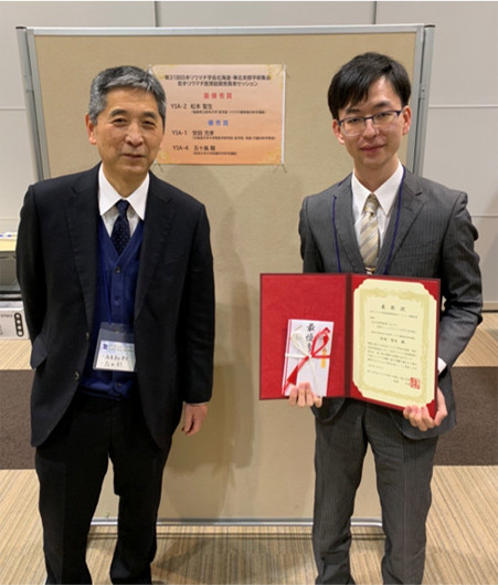 松本聖生先生が第31回日本リウマチ学会で最優秀賞を受賞し、賞状を持って笑顔でポーズをとる様子。背景には受賞セッションの掲示が見える。