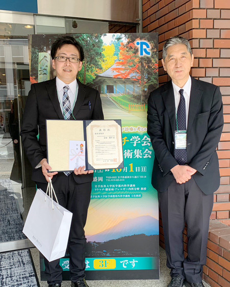 吉田周平先生が第33回日本リウマチ学会で優秀演題賞を受賞し、賞状を持っている写真。背景には学会のポスターが見える。