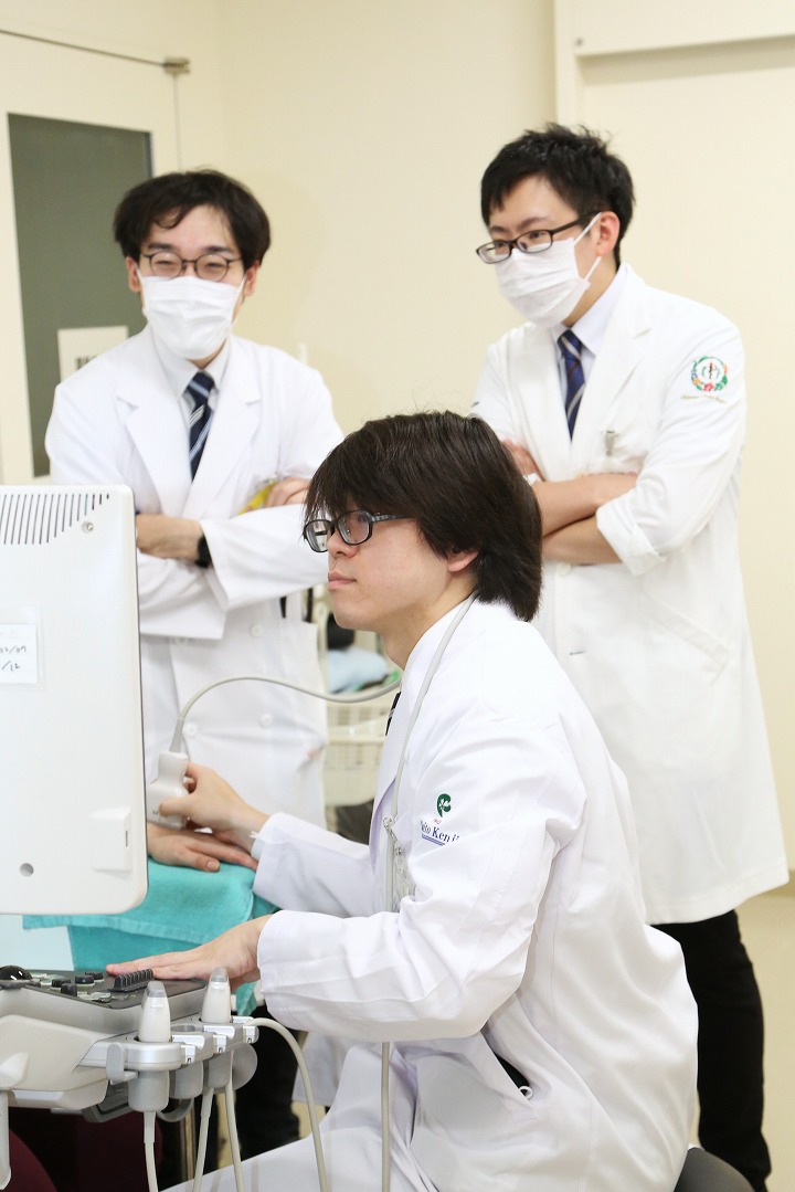 白衣を着た3人の医療従事者が医療用モニターを見つめて議論している。主に画面を操作しているのは、椅子に座っている若い男性。