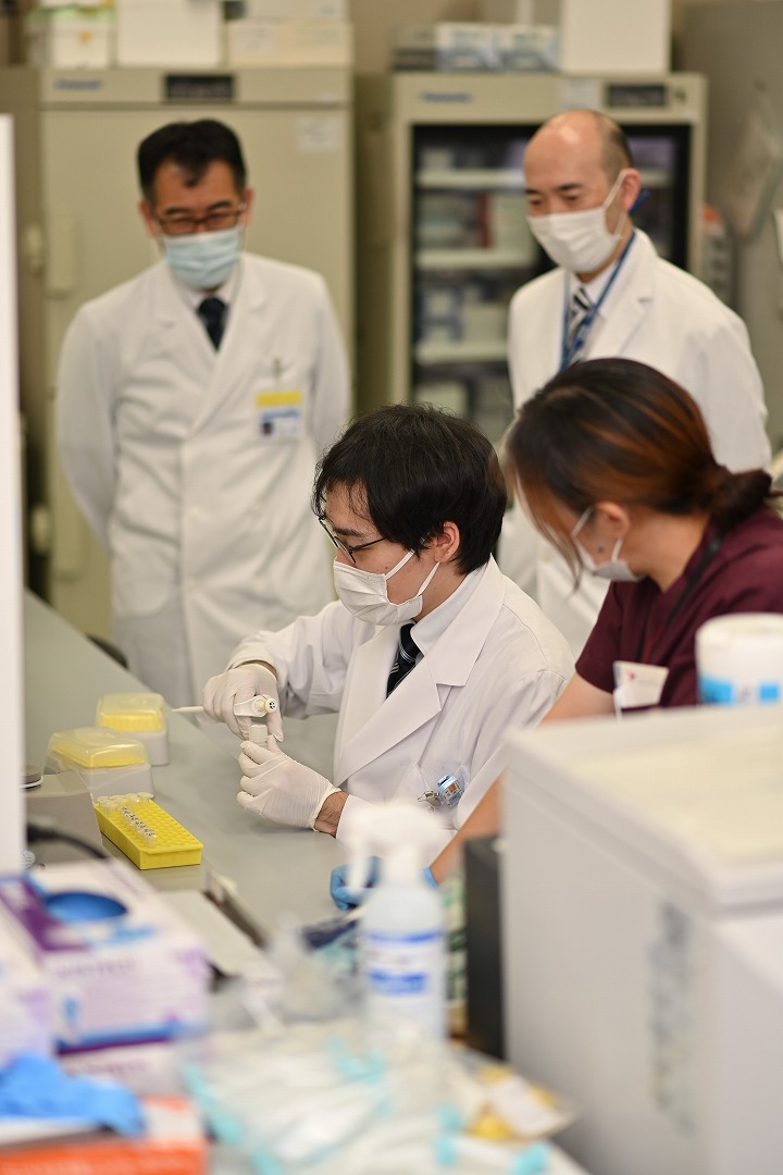 研究室で実験に取り組む白衣姿の学生たち。周囲には研究機材が整然と配置されており、真剣な表情で観察している。