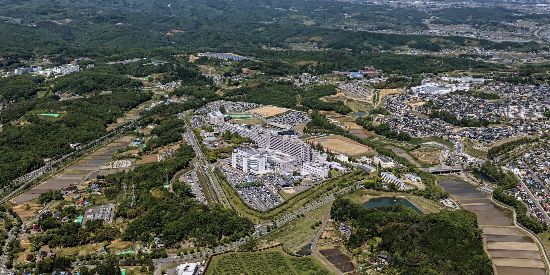福島県立医科大学のキャンパスの空撮画像。広範囲に広がる施設や駐車場、周囲の自然豊かな風景が見える。