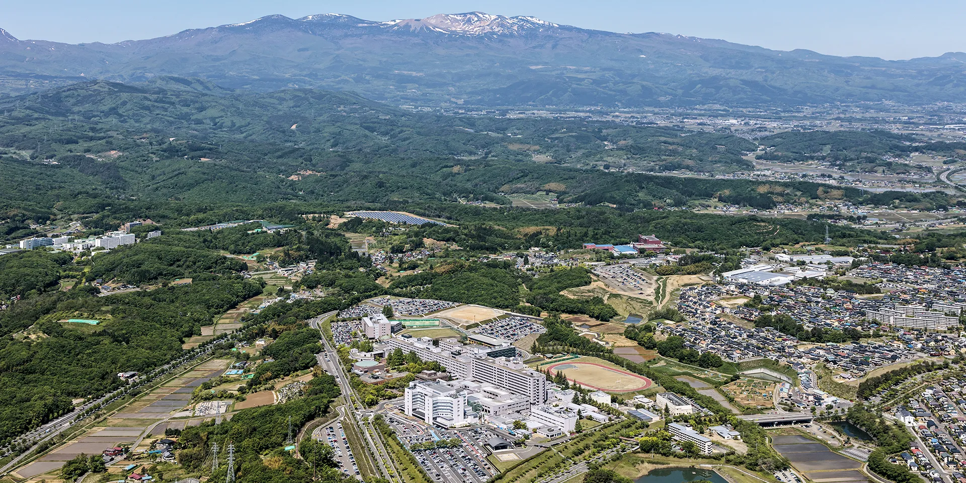 福島県立医科大学のキャンパスの空撮画像。広範囲に広がる施設や駐車場、周囲の自然豊かな風景が見える。