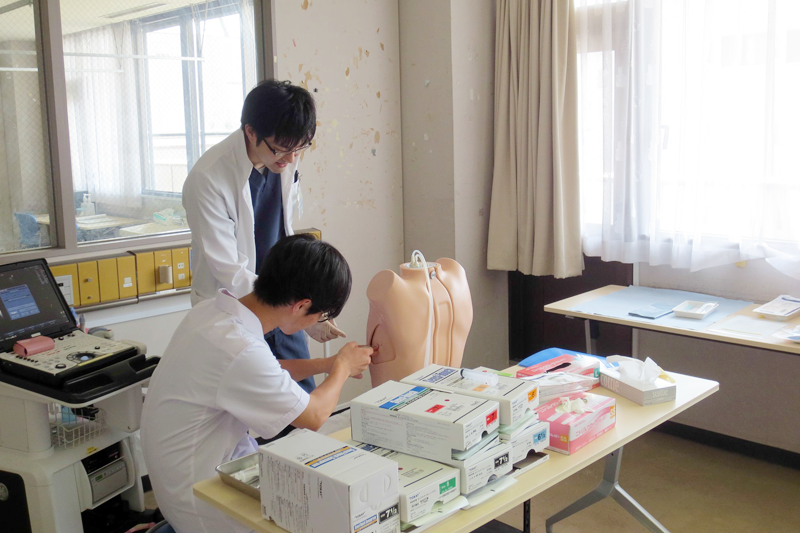 医療研修中の学生と指導者が模型を使い、臨床手技の練習を行っています。机には医療器具が並んでいます。