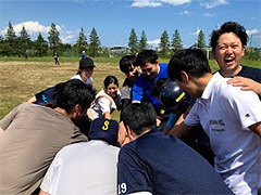 グループの人々がアウトドアで円を作り、笑顔で手を重ね合わせている。青空と木々が背景。