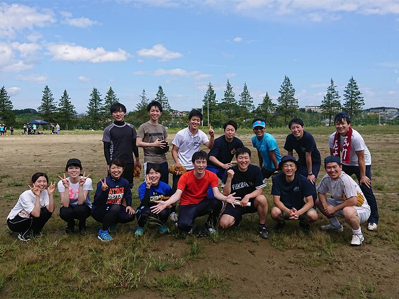 野外のフィールドで、スポーツウェアを着た学生たちが笑顔で集まり、グループ写真を撮っています。青空と木々が背景に広がっています。
