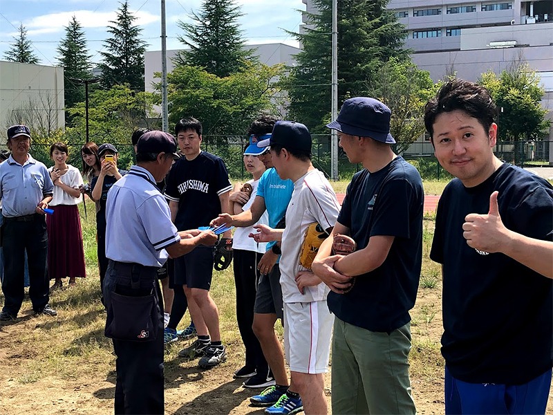 屋外で集合した人々が並び、一人が紙を受け取っている。手前の男性が親指を立ててポーズ。背景は木々と建物。