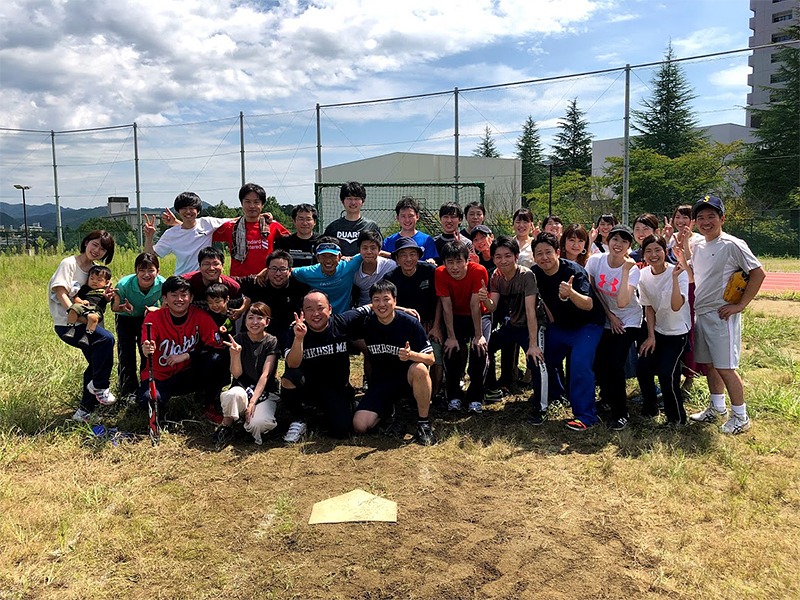 大勢の大学生たちが野外で楽しそうに集まり、スポーツイベントに参加している様子。背景に青空と木が見える。
