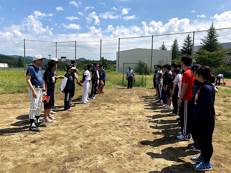 野外のグラウンドで対面して整列する2列の人々。みなスポーツウェアを着用し、晴天の下で何かの競技前ミーティングを行っている。後方には建物と青空が広がる。