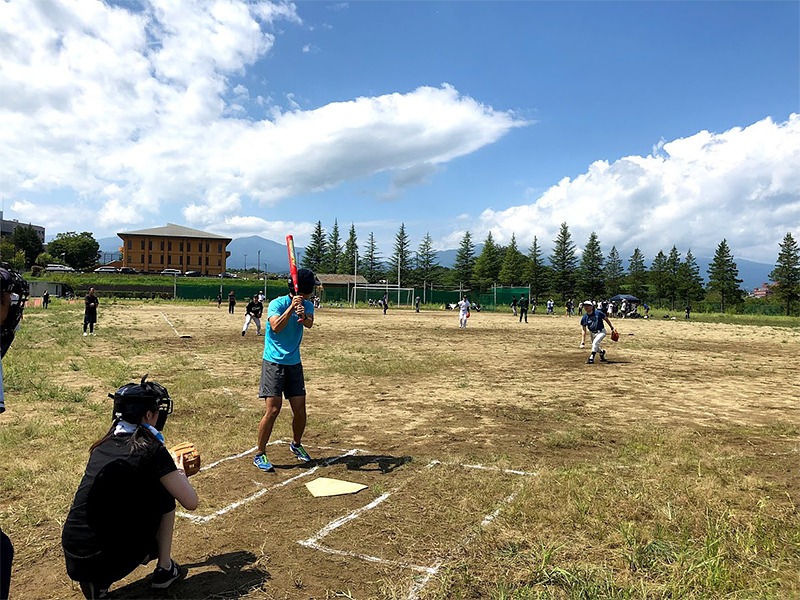 野外の野球グラウンドで、バッターがバットを構え、キャッチャーがその後ろに座っています。背景には青空と木々、建物が見えます。