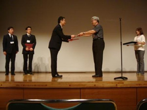 表彰式で男性が壇上で賞状を受け取っている様子