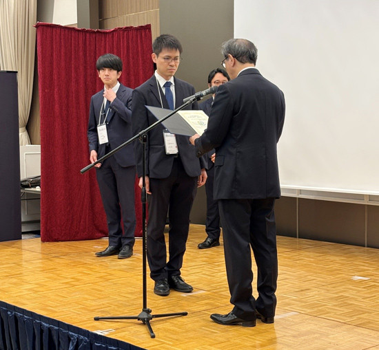 壇上でスーツ姿の男性が証書を受け取っている様子。