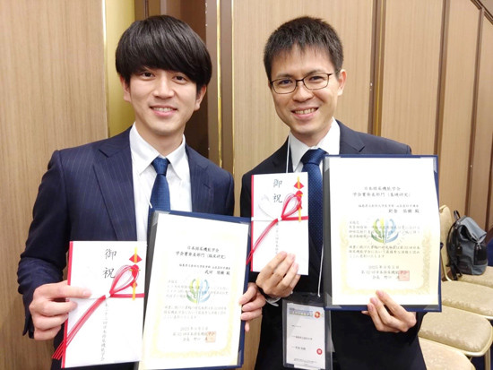 スーツ姿の男性二人が賞状を持って笑顔で立っている。