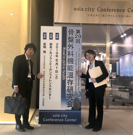 2人の人物が「第29回骨盤外科機能温存研究会」の看板の前に立っている様子。