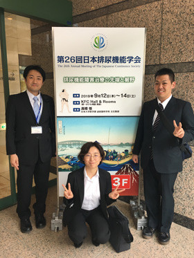 3人のビジネススーツ姿の人物が、第26回日本排尿機能学会の看板の前に立っている様子。