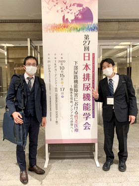 二人の男性が、第27回日本排尿機能学会の大きな看板の前でマスクを着用して立っている様子。